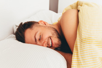 Fototapeta na wymiar A young european smiling man with beard is waking up in a bed on pillows with nice mood. Good morning