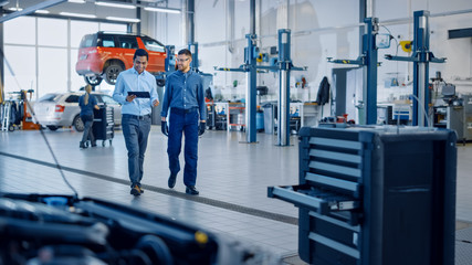 Manager Checks Diagnostics Results on a Tablet Computer and Explains a Vehicle Breakdown to a Handsome Mechanic. Car Service Employees Talk while Walking in a Garage. Modern Clean Workshop.