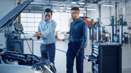 Fototapeta premium Manager Checks Data on a Tablet and Explains an Engine Breakdown to a Handsome Professional Mechanic. Car Service Employees Inspect a Car with Internal Combustion Engine. Modern Clean Workshop.