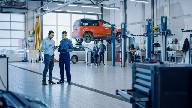 Manager Checks Diagnostics Results on a Tablet Computer and Explains a Vehicle Breakdown to a Handsome Mechanic. Car Service Employees Talk while Standing in a Garage. Modern Clean Workshop.