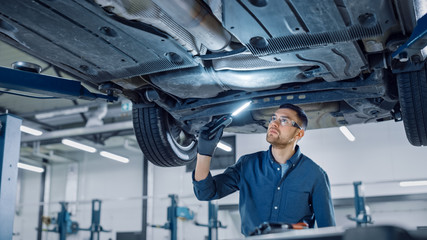 Handsome Professional Car Mechanic is Investigating Rust Under a Vehicle on a Lift in Service. Repairman is Using a LED lamp and Walks Towards. Specialist is Wearing Safety Glasses. Modern Workshop. - obrazy, fototapety, plakaty