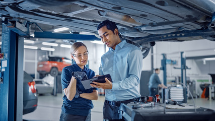 Female Mechanic Talking to a Manager Under a Vehicle in a Car Service. Specialist is Showing Info on a Tablet Computer. Empowering Woman Wearing Gloves and Safety Gloves. Modern Clean Workshop.  - Powered by Adobe