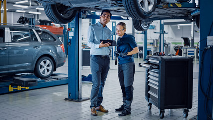 Female Mechanic Talking to a Manager Under a Vehicle in a Car Service. Specialist is Showing Info on a Tablet Computer. Empowering Woman Wearing Gloves and Safety Gloves. Modern Clean Workshop. 