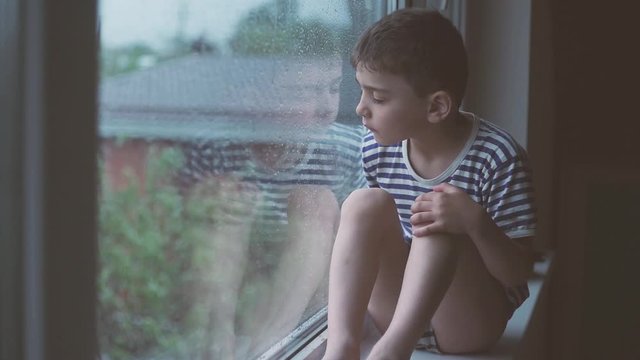 a little boy sits outside a rainy window and is sad. A window in rainy weather behind which a small sad boy sits, a child is hard going through self-isolation, orphaned children in orphanages and boar