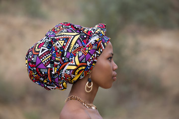 Profile portrait of a young dark-skinned African girl in a national headdress and gold jewelry - obrazy, fototapety, plakaty