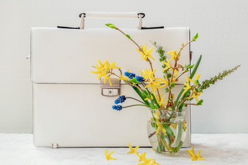White briefcase, and bouquet of blue and yellow flowers in vase close-up, selective focus. Concept of spring or summer shopping, back to school