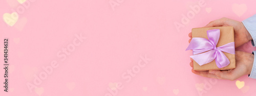 Kid's hands holding gift box wrapped in craft paper and tied with bow on pink background. Concept Mother's Day banner.