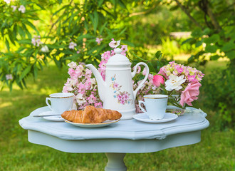 Coffee break in blooming summer garden. Vintage coffee pot, two cups of coffee, croissant and bunch of rose flowers on vintage table in the garden