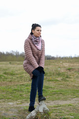 Naklejka na ściany i meble Girl in the spring meadow. He stands with his foot on a stone. Dressed in a spring jacket, a scarf around her neck.