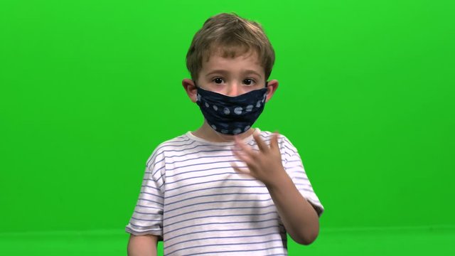 Little child with reusable cloth mask signs "Thumbs  up" and takes off his mask over green screen.