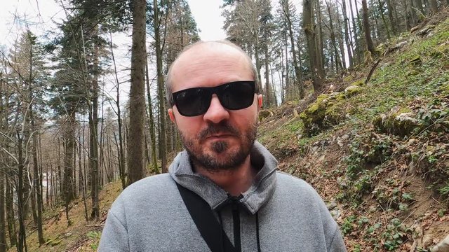 Adult man in hoodie and sunglasses walking on wood trail and recording video blog with action camera
