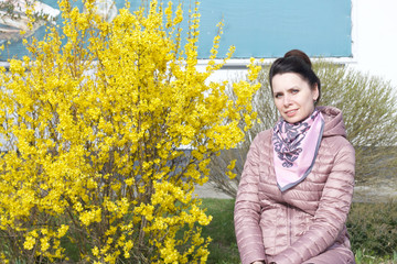 Portrait of a smiling girl. Against the background of a bush strewn with yellow flowers. Dressed in a spring jacket, a scarf around her neck.