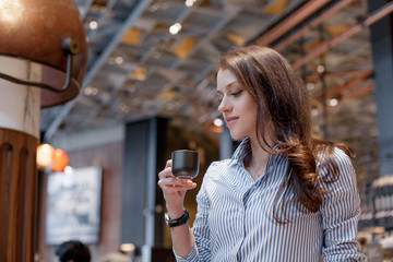 Young beautiful girl in a cafe looks at a mug with coffee in a black cup. Girl in a fashionable blue shirt