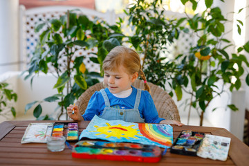 little toddler girl painting rainbow and sun with water colors during pandemic coronavirus quarantine disease. Children painting rainbows around the world with the words Let's all be well. Happy child