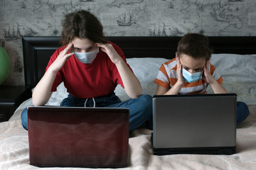 schoolboy and schoolgirl in medical masks sit on bed in their room, look in laptop screens, do their homework, remote education concept, stay home during coronavirus pandemic