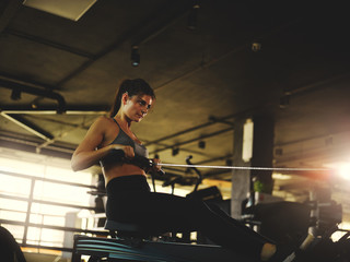Fototapeta na wymiar Side view backlit shot of determined strong woman doing exercise on rope pull machine in gym