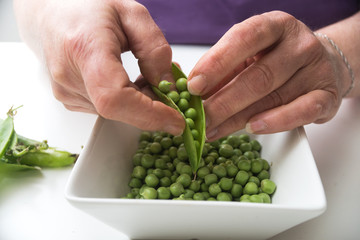 Naklejka na ściany i meble Closeup of hands of woman peeling fresh organic peas on white table background