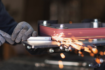 Industrial tool worker grinds a square steel pipe on a rotating belt sander - obrazy, fototapety, plakaty