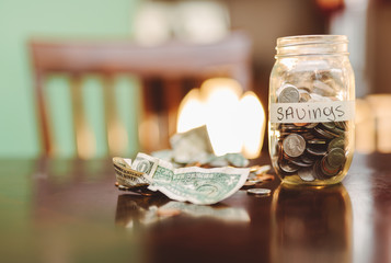 mason jar filled with coins along with US dollar bills on brown table with light in background