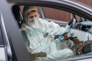 Man in protective suit, mask and gloves washing and disinfection of the steering wheel in the car, prevent infection of Covid-19 virus, contamination of germs or bacteria. Concept coronavirus pandemic