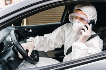 Man in protective suit, medical mask and rubber gloves for protect from bacteria and virus is sitting in a car and talking on the phone. Concept protection during the covid 19 coronavirus pandemic.