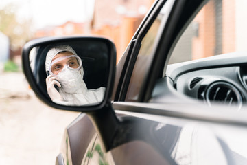 Man in protective suit, medical mask and  gloves is driving a car and talking on the mobile phone. Concept of protection against coronavirus infection in the context of the global pandemic covid 19.
