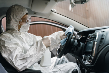 Man in protective suit, mask and gloves washing and disinfection of the steering wheel in the car, prevent infection of Covid-19 virus, contamination of germs or bacteria. Concept coronavirus pandemic