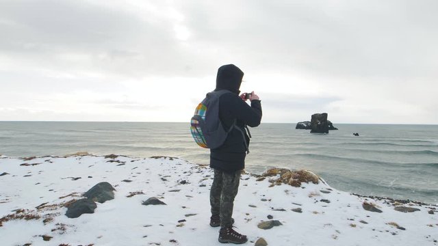 Young man tourist takes pictures at the top of the cliff around Reynisfjara Beach and Dyrholaey lighthouse in Iceland. South part of Iceland close to Vik at winter
