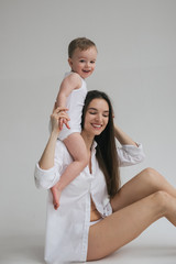 Young beautiful brunette mom plays with her little baby son. Stylishly dressed in white clothes. Hug, happy together, play. Studio portrait. Motherhood concept, tender feelings.