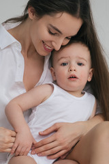 Young beautiful brunette mom plays with her little baby son. Stylishly dressed in white clothes. Hug, happy together, play. Studio portrait. Motherhood concept, tender feelings.