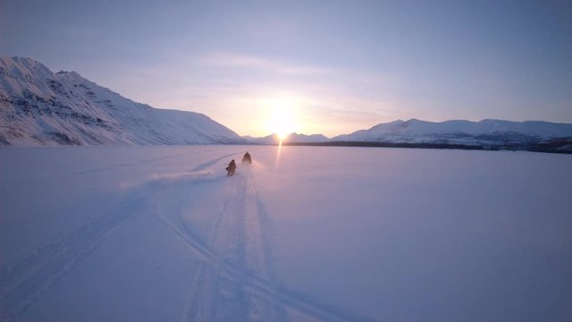 Skier riding behind snowmobile on snowy plain on evening sunset landscape in mountain. Men riding ski behind snowmobile on snowy field in mountain terrain on colorful sunset background.