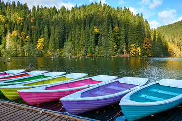 Fototapeta premium Multi-colored boats at the pier on Lake Lacu Rosu, Romania. Autumn landscape of mountain lake, forest and mountains.