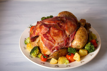 Whole roast chicken with breasts wrapped in bacon and vegetables: potatoes, broccoli and tomatoes. horizontal image
