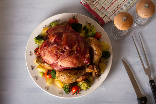 Whole roast chicken with breasts wrapped in bacon and vegetables: potatoes, broccoli and tomatoes. Overhead horizontal image