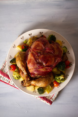 Whole roast chicken with breasts wrapped in bacon and vegetables: potatoes, broccoli and tomatoes. Overhead vertical image