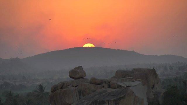 Flocks of birds slowly fly against the background of sunset or dawn, in the valley above the large rocks in India, Hampi. Slow motion.