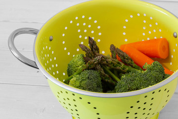 Washed asparagus, broccoli and carrot vegetables in a bright green colander strainer.  With white wood background