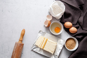 Baking cooking ingredients: a bag of flour, eggs, egg with 
yolk in a bowl,  brown sugar, butter dark linen cloth, rolling pin, on light grey concrete background. Top View Copy Space.  Recipe Mockup