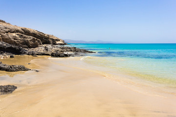 Fototapeta na wymiar Crystal clear water beach by volcanic rocks coast on sunny day in Fuerteventura. Calm, transparent sea ideal for relax and peaceful vacation. Summer holidays, tourism travel destination concepts