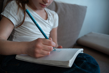 Kids home Quarantine.Emotional portrait of a smiling girl sitting at a desk and drawing on paper. Girl listens to music on headphones. The light of the setting sun. Close-up