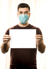 Theme of coronavirus and safety. A young man in a mask from a virus stands with a white sheet for a mockup, on a light background. Vertical frame