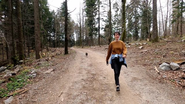 Woman in casual clothes walking with small black dog on the forest macadam trail, footage with action camera