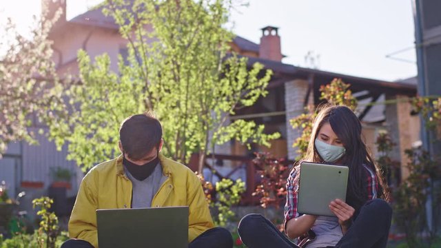 Sunny day at home in the garden attractive lady and man sitting in the grass while is qua they studying from home using tablet and laptop they wearing protective mask new Covid-19 pandemic