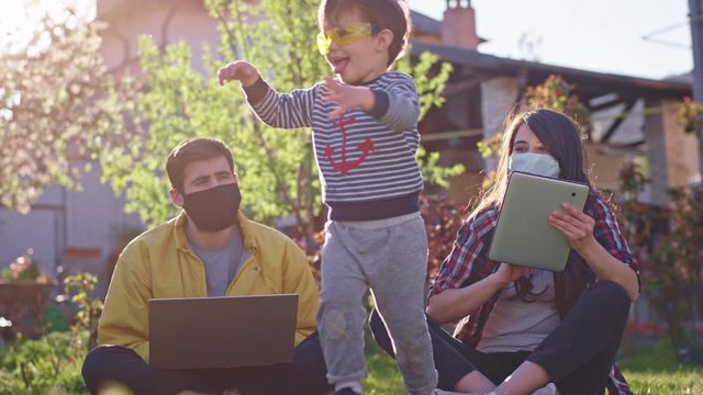 At home mom and dad working in the garden while sitting down on the grass they using the laptop and tablet to work online wearing protective mask their small son playing around them