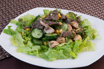 Salad with pieces of meat, mushrooms, cucumbers, pickled corn, dill, lettuce, olive oil on a large square white plate on a brown background
