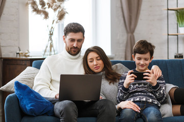 Mother, father and son using laptop and mobile phones at home. Family members ignoring each other and live talk. Gadget influence on family relationships. Modern technologies and addiction concept.