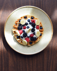 Belgian waffle with bananas, strawberries, blackberries, blueberries and whipped cream from above on wood table with copy space