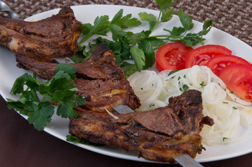 Roasted lamb ribs with fat on coals on a stick on a white oval dish with parsley, tomatoes, onion rings