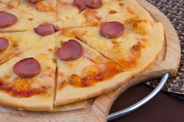 Big cheese pizza with tomatoes and smoked sausage on a wooden round board close-up on a brown background