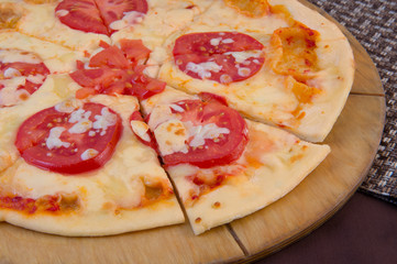Cheese pizza with large tomatoes on a wooden round plate on a brown background